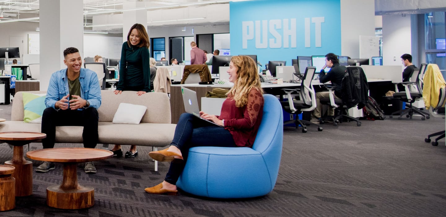 Membros da equipe diversa do Sprout Social sentados em móveis bonitos em frente a um quadro com a frase "Push it" feita de pinos.